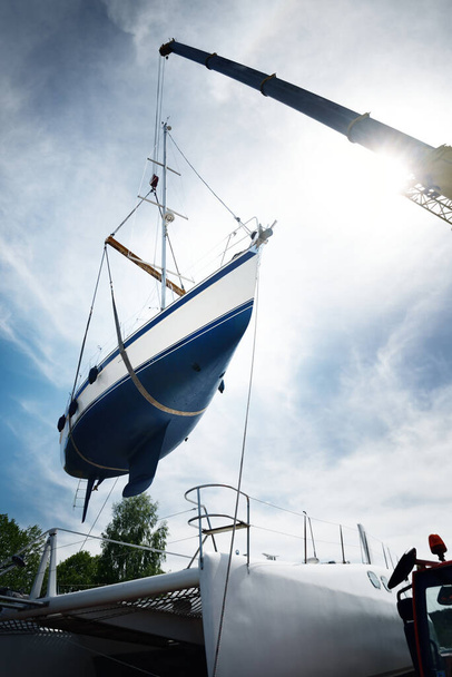 Blaues Segelboot, angehoben von einem Kran, der nach der Überwinterung im Winter wieder ins Wasser gesetzt wird. Klarer blauer Himmel mit Zirruswolken. Hafen von Riga, Lettland. Thema Transport, Sport und Erholung - Foto, Bild