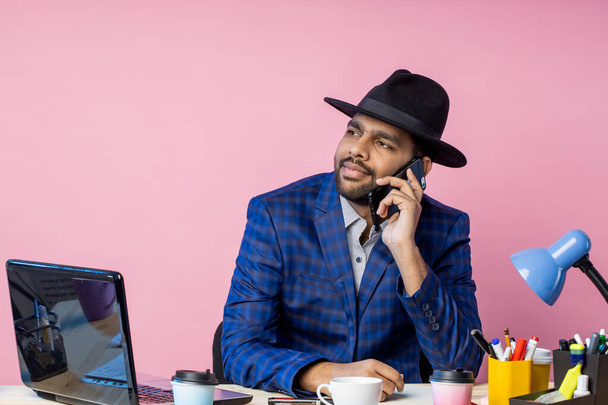 Joyful stylish indian entrepreneur wearing formal wear, black hat, talking on mobile phone, smiling broadly, looking to side, sitting at desk in office against pink wall . Job, business concept. - Фото, изображение