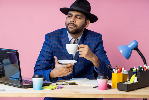 young unshaven indian businessman in formal blue checkered suit, black hat , sitting at workplace, thinking about something, looking aside, enjoying aromatic coffee or tea. - Zdjęcie, obraz