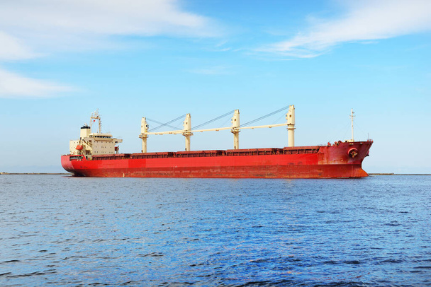Duży czerwony masowiec (statek towarowy) z dźwigami pływającymi na otwartym morzu z Europoort. Czyste błękitne niebo z chmurami cyrru. Rotterdam, Holandia. Komunikacja globalna, logistyka, temat przemysłu - Zdjęcie, obraz