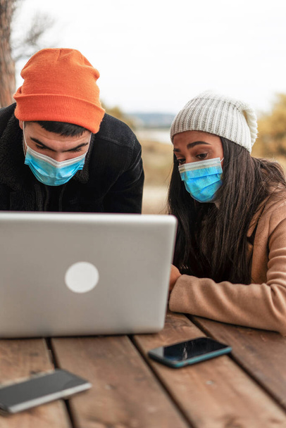 Молодая многонациональная пара друзей, использующих ноутбук на открытом воздухе в защитной маске и зимней одежде. Латиноамериканка, сидящая за деревянным столом в сосновом лесу, просматривает интернет во время зимних каникул - Фото, изображение