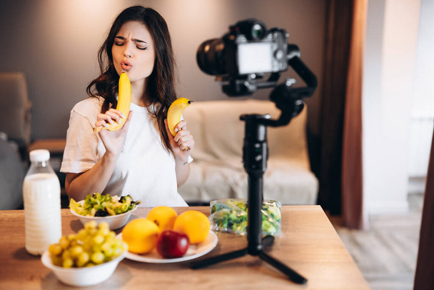 健康食品ブロガー若い女性は、キッチンスタジオで果物ビーガンサラダの新鮮な料理,ビデオチャンネルのためのカメラでチュートリアルを撮影.女性のインフルエンサーはジャンクフードを見せず、健康的な食事について話します. - 写真・画像