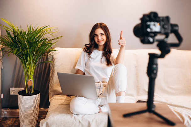 Молодая удивленная блоггерша сидит на диване с ноутбуком, записывая свой видеоблог речи перед аудиторией. Блогинг внутри. Прямая трансляция. - Фото, изображение