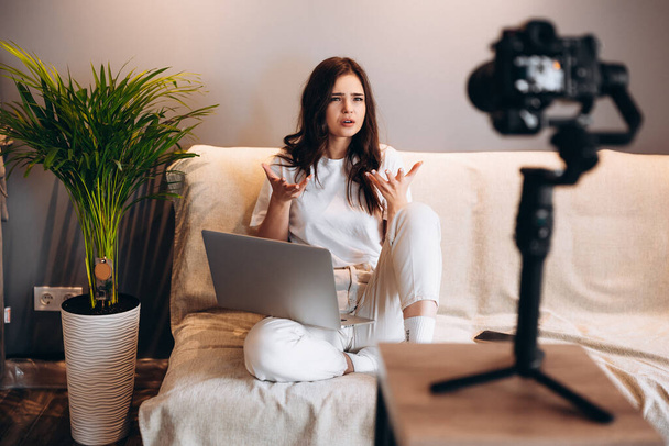 Giovane donna blogger sorpresa è seduta sul divano con computer portatile registrando il suo vlog discorso al suo pubblico. Blogging indoor. - Foto, immagini