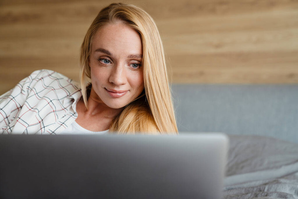 Beautiful blonde smiling woman using laptop while lying on bed at home - Zdjęcie, obraz