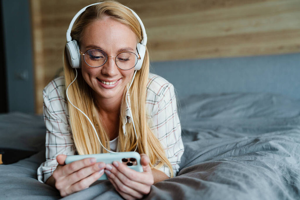 Blonde smiling woman listening music with earphones and mobile phone on bed at home - Photo, image