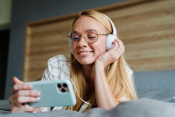 Blonde smiling woman listening music with earphones and mobile phone on bed at home - Fotoğraf, Görsel