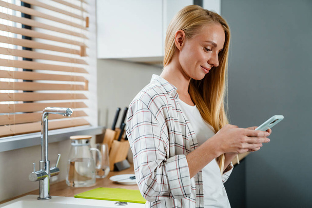 Blonde happy woman using mobile phone and smiling at home kitchen - Fotoğraf, Görsel