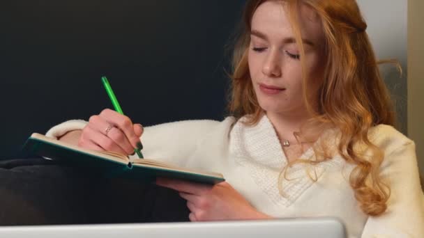 Mujer caucásica con el pelo rizado rojo está escribiendo algo en un copybook sonriendo mientras el trabajo en el ordenador portátil - Imágenes, Vídeo