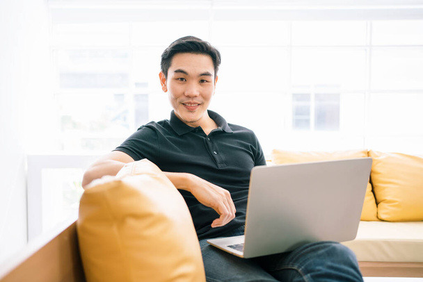 アジアの男性は、ラップトップで作業しながらカメラを見て、ソファに座って笑っている。自宅のノートパソコンで働くカジュアルなビジネスマン。ビジネスにおける問題解決のプロセス. - 写真・画像