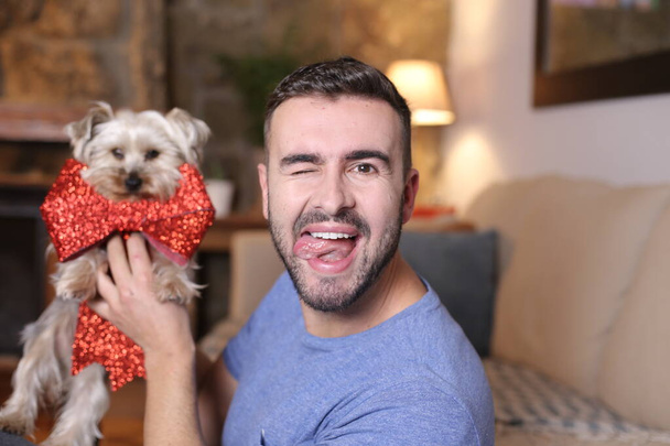 retrato de joven guapo sosteniendo perro con lazo decorativo rojo en casa - Foto, Imagen