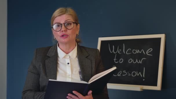 Maestra mayor caucásica con gafas está explicando la lección cerca de la pizarra sosteniendo un libro y hablando con los estudiantes - Metraje, vídeo