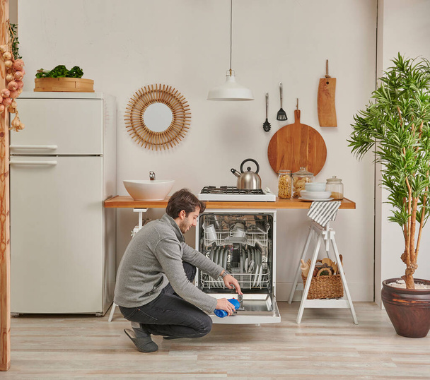 Ο άνθρωπος γεμίζει υγρό απορρυπαντικό στο πλυντήριο πιάτων, διακοσμητική μικρή κουζίνα, νεροχύτη και ψυγείο. - Φωτογραφία, εικόνα