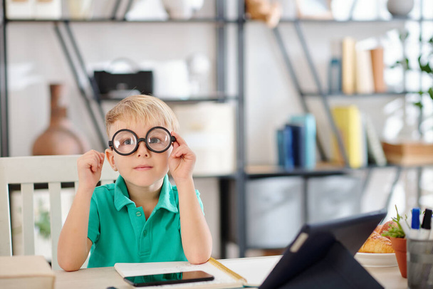 Funny schoolboy putting on glasses with thick lenses and looking at camera when sitting at his desk at home - Photo, Image