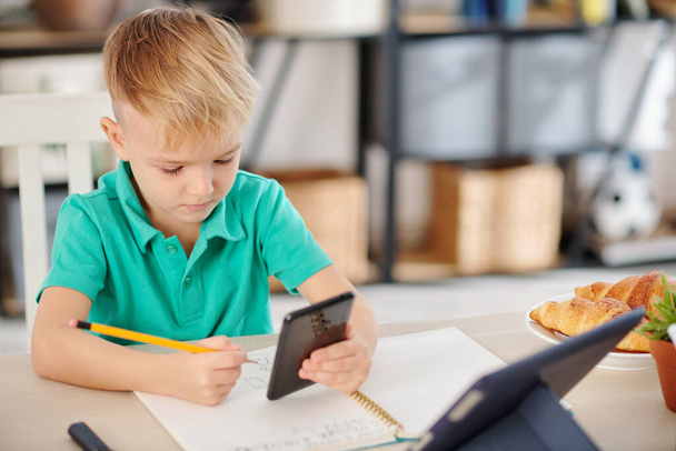 Serieuze schooljongen controleren van informatie op smartphone bij het oplossen van wiskunde vergelijkingen aan zijn bureau - Foto, afbeelding