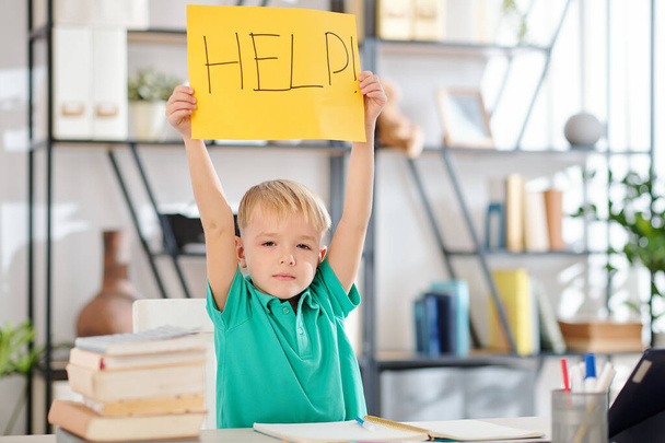 Portrait of preteen schoolboy sitting at table, holding help sign over his head and looking at camera - Photo, Image