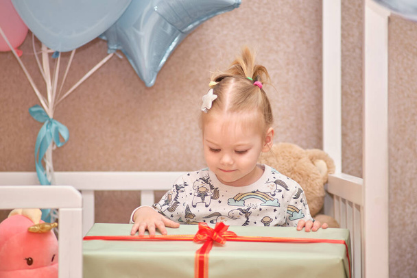 Una niña pequeña en pijama recibe regalos de cumpleaños mientras está sentada en una cuna blanca. Globos en el fondo - Foto, imagen