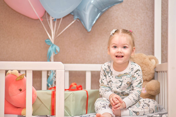 Ένα κοριτσάκι με πιτζάμες λαμβάνει δώρα γενεθλίων ενώ κάθεται σε ένα λευκό βρεφικό κρεβατάκι. Μπαλόνια στο παρασκήνιο - Φωτογραφία, εικόνα