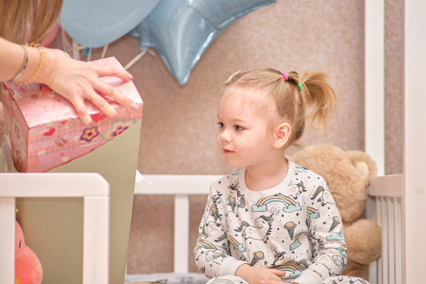 Une petite fille en pyjama reçoit des cadeaux d'anniversaire alors qu'elle est assise dans un lit bébé blanc. Ballons en arrière-plan - Photo, image