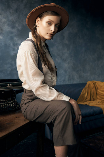portrait photo of a girl in retro style posing in an interior studio wearing pants, shirt, suspenders and hat, she sits on the table and looks at camera - Photo, Image