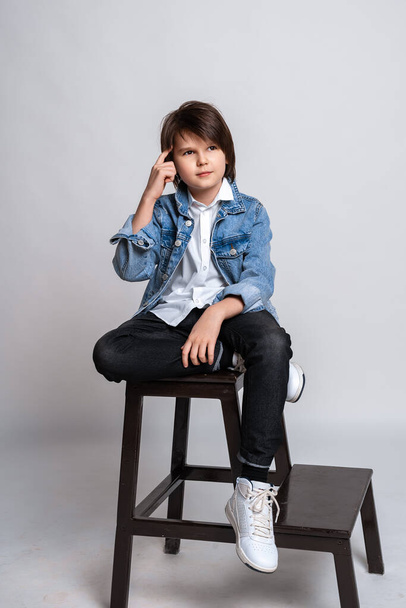 Przystojny chłopiec w modzie 11-13 lat siedzi na krześle w studio. Ubrany w białą koszulę, czarne dżinsy, dżinsową kurtkę i białe trampki. Test modelowy. Koncepcja mody i ludzi - Zdjęcie, obraz