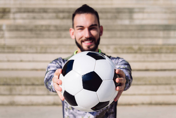 football ball in the hands of a smiling sportsman in a concrete soccer court, selective focus on the ball, concept of healthy lifestyle and urban sport in the city, copy space for text - Фото, изображение