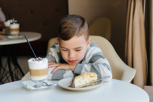 A cute little boy is sitting in a cafe and looking at a cake and cocoa close-up. Diet and proper nutrition - Foto, afbeelding