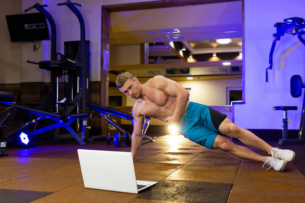 Αθλητής ξανθός άνδρας εκτελεί ασκήσεις γυμναστικής στο γυμναστήριο και προπονείται online με φορητό υπολογιστή - Φωτογραφία, εικόνα