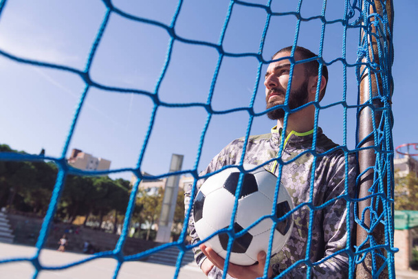 portrait of an attractive sportsman through the goal net with the football ball in his hand, concept of healthy lifestyle and urban sport in the city, copy space for text - Fotografie, Obrázek