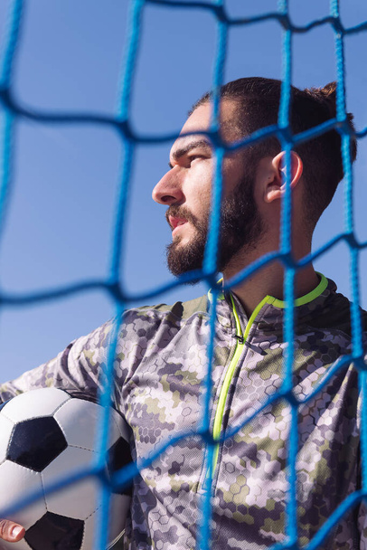 vertical portrait of an attractive sportsman through the goal net with the football ball under his arm, concept of healthy lifestyle and urban sport in the city - Photo, Image