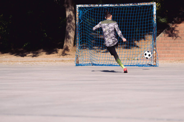 rear view of an unrecognizable sportsman shooting the soccer ball to an empty goal in a concrete football court, concept of healthy lifestyle and urban sport in the city, copy space for text - Photo, Image