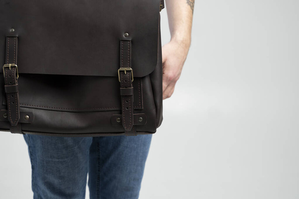 Details of brown mens shoulder leather bag for a documents and laptop on the shoulders of a man in a blue shirt and jeans with a white background. Satchel, mens leather handmade briefcase. - Fotografie, Obrázek
