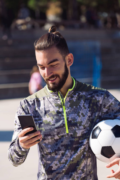 fashionable sportsman with a soccer ball smiles consulting his mobile phone on the football court, concept of technology and urban sport lifestyle in the city - Fotografie, Obrázek