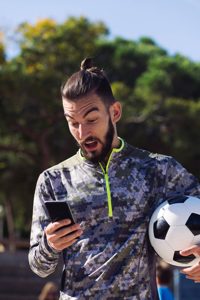 fashionable sportsman with a soccer ball consulting his mobile phone with surprised expression on the football court, concept of technology and urban sport lifestyle in the city - Photo, Image