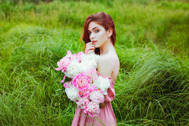 ピンクの牡丹の大きな花束と自然の中で若い赤毛の女性の屋外肖像画。女の日だ。女性の春、夏のファッションコンセプト. - 写真・画像