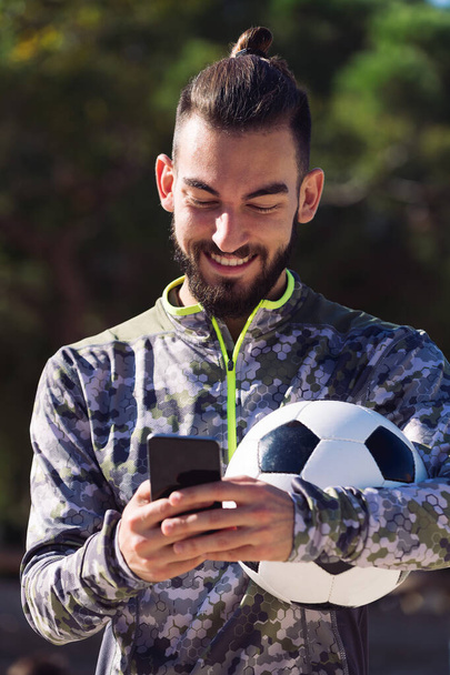 portrait of a happy sportsman with a soccer ball smiles writing a message on his mobile phone on the football court, concept of technology and urban sport lifestyle in the city - Foto, Imagem