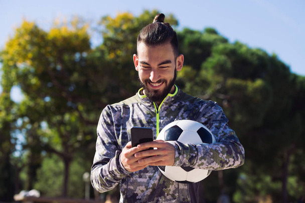 happy sportsman with a soccer ball smiles writing a message on his mobile phone, concept of technology and urban sport lifestyle in the city, copy space for text - Photo, Image
