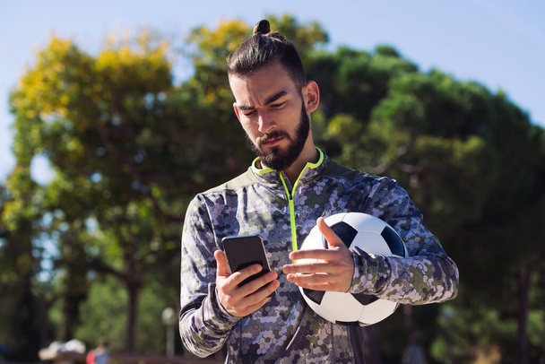 fashionable sportsman with a soccer ball consulting his mobile phone with confused expression due to an unexpected message, concept of technology and urban sport lifestyle in the city - Photo, Image