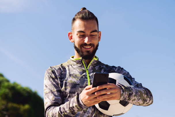 horizontal portrait of a happy sportsman with a soccer ball smiling writing a message on his mobile phone, concept of technology and urban sport lifestyle in the city, copy space for text - Photo, Image