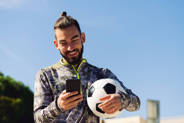 horizontal portrait of a happy sportsman with a soccer ball smiling looking his mobile phone, concept of technology and urban sport lifestyle in the city, copy space for text - Photo, image