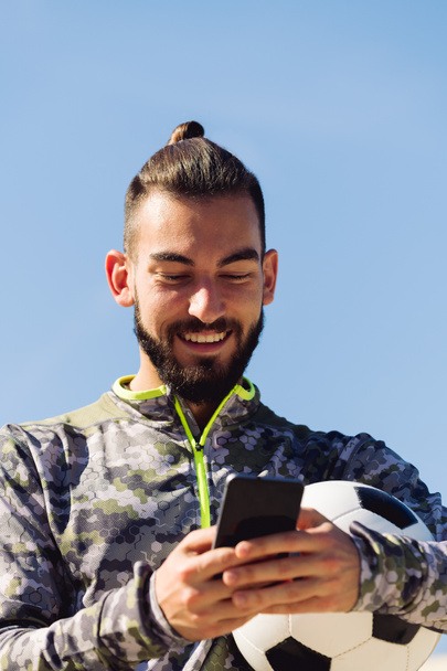 vertical portrait of a happy sportsman with a soccer ball laughing looking his cell phone, concept of technology and urban sport lifestyle in the city, copy space for text - Foto, afbeelding
