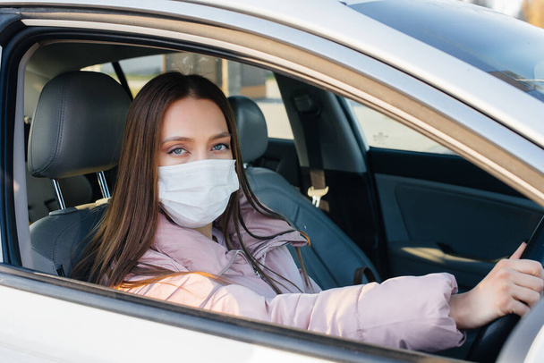 A young girl sits behind the wheel in the car in the mask during the global pandemic and coronavirus. Quarantine - Fotoğraf, Görsel