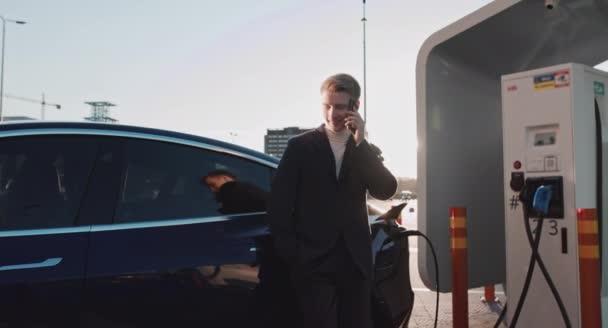 Uomo d'affari caucasico che utilizza smartphone e si appoggia sulla lussuosa auto elettrica blu che sta caricando. Spina di ricarica di un'auto elettrica. Carica auto elettrica di lusso. Uomo d'affari biondo parlando al telefono - Filmati, video