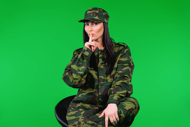 Serious young woman in military uniform shows quietly tsss with finger to the mouth, looking at the camera. Green background. - Фото, зображення