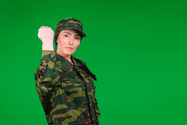 Young woman in military uniform shows her fist to the camera on a green background with blank space. Strong fearless female soldier - Foto, afbeelding
