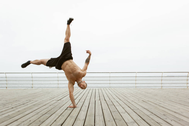 Μυώδης bodybuilder fitness άνθρωπος με γυμνό κορμό στέκεται στο ένα χέρι στην παραλία - Φωτογραφία, εικόνα