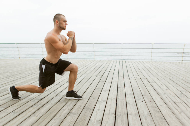atleta masculino muscular com tronco nu fazendo lunges exercício na praia - Foto, Imagem