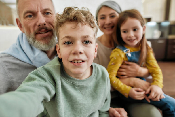 Excited little boy making a selfie with family at home, cheerful grandparent spending time with grandchildren, playing and having fun in the living room - Фото, изображение