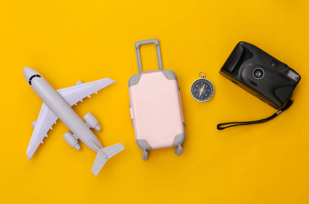 Podróżować płasko. Mini plastikowa walizka podróżna, aparat fotograficzny, samolot i kompas na żółtym tle. Minimalny styl. Widok z góry - Zdjęcie, obraz