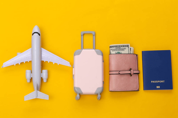 Die Reise lag flach. Mini-Reisekoffer aus Kunststoff, Geldbörse, Flugzeug und Reisepass auf gelbem Hintergrund. Minimaler Stil. Ansicht von oben - Foto, Bild